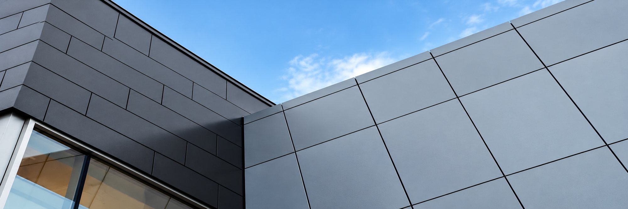 Angle d’un bâtiment contemporain habillé de panneaux de bardage gris foncé, avec une fenêtre en bas à gauche et un ciel bleu clair au fond.