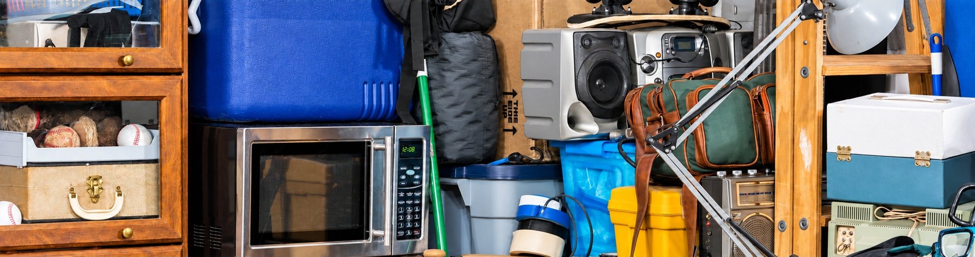 Un espace de stockage rempli d’objets entassés : un four à micro-ondes, des radios, une glacière bleue, des vitrines en bois et des valises.
