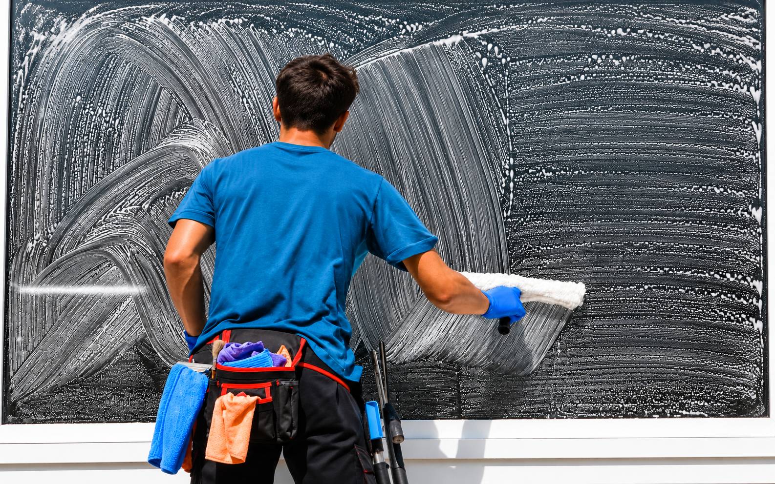 Un professionnel en t-shirt bleu et gants bleus, vu de dos, lave une grande vitre savonneuse avec un applicateur ; une ceinture porte-outils à la taille.